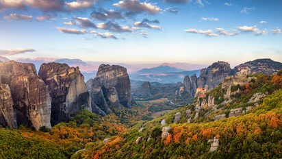 Man blickt über eine urwüchsige, gebirgige Landschaft. Zwischen kargen aufragenden Felskomplexen überziehen grüne, schon leicht herbstlich gefärbte Wälder die Landschaft. Rechts im Bildmittelgrund erkennt man auf einigen Anhöhen die berühmten Meteora-Klöster.