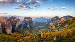 Man blickt über eine urwüchsige, gebirgige Landschaft. Zwischen kargen aufragenden Felskomplexen überziehen grüne, schon leicht herbstlich gefärbte Wälder die Landschaft. Rechts im Bildmittelgrund erkennt man auf einigen Anhöhen die berühmten Meteora-Klöster.