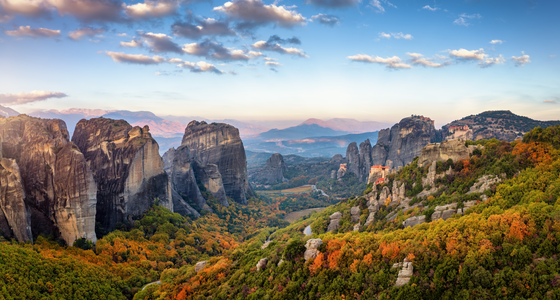 Landschaft in Epirus mit den Meteora-Klöstern Man blickt über eine urwüchsige, gebirgige Landschaft. Zwischen kargen aufragenden Felskomplexen überziehen grüne, schon leicht herbstlich gefärbte Wälder die Landschaft. Rechts im Bildmittelgrund erkennt man auf einigen Anhöhen die berühmten Meteora-Klöster.
