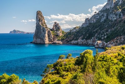 Aus dem leuchtend-blauen Meer erhebt sich in der Bildmitte eine Felsnadel. Nach rechts steigt ein Berg aus dem Meer auf. Der Bildvordergrund wird von grüner Vegetation eingenommen.