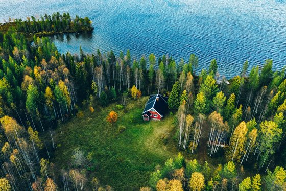 Finnische Sommerfrische, das Mökki Man schaut aus der Luft auf eine Wald- und Wasserlandschaft, in der auf einem Wiesenteil ein rotes Häuschen steht.