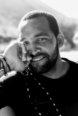 Schwarz-Weiß-Porträt eines etwa 40-jährigen Kapverdiers mit kurzem krausen Haar und Vollbart, der direkt in die Kamera schaut und seinen Kopf auf die rechte Hand stützt.. Er trägt ein schwarzes T-Shirt und Armbänder um sein Handgelenk.