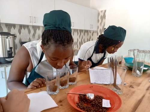 Qualitätskontrolle in der Kooperative ©Angelique's Finest Zwei Women of Colour mit weißen Oberteilen und schwarzen Schürzen riechen an je drei mit Kaffeepulver gefüllten Gläsern in einer modernen Küche.