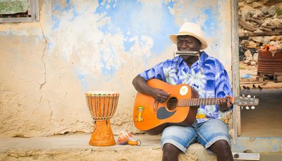 Kapverdischer Musiker sitzt vor einer weiß-blauen Wand und spielt Akustikgitarre und Mundharmonika, neben ihm eine hölzerne Trommel.  