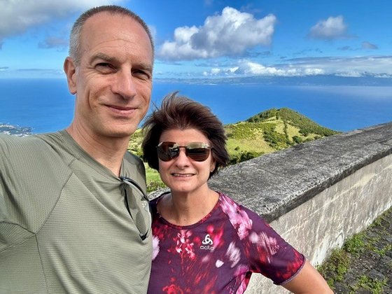 Selfie eines mittelalten Hetero-Paares, das die linke Bildhälfte einnimmt. Der Mann steht am linken Bildrand, die frau rechts neben ihm. Im Hintergrund sieht man eine Mauer und dahinter in der Tiefe blaues Meer.