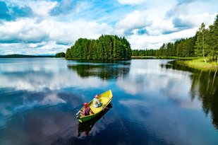 In einem Boot auf einer großen Wasserfläche sitzt ein angelndes Paar, am Ufer im Hintergrund stehen Bäume.