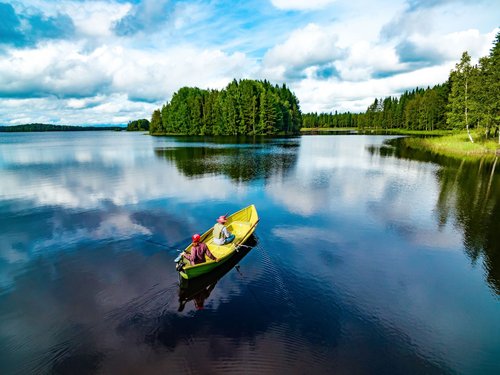 In einem Boot auf einer großen Wasserfläche sitzt ein angelndes Paar, am Ufer im Hintergrund stehen Bäume.