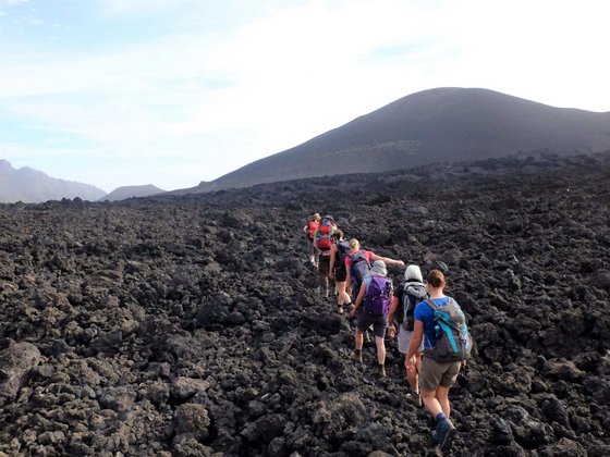 In einer schwarzen Lavalandschaft bewegt sich eine Gruppe Wanderer von rechts unten in den Bildmittegrund hinein. Rechts hinten erhebt sich ein Berg, darüber blau-weißer Himmel.