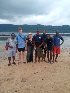 Eine Gruppe von Menschen stehen am Strand im Sand, hinter ihnen sieht man das Meer und dahinter Hügel. Die Gruppe besteht aus einer europäischen Frau und einem ebenfalls europäischen Jugendlichen sowie fünf afrikanischen Männern.