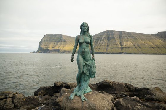 Einer Legende entsprungen: die Robbenfrau Die patinierte Bronzeskulptur einer Frau mit einer Robbenhaut über ihrem linken Bein steht vor einer Küstenkulisse.