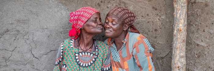 Zwei ältere Damen sitzen entspannt vor einer bemalten Hausmauer in einem Dorf in Afrika und lächeln einander zu.