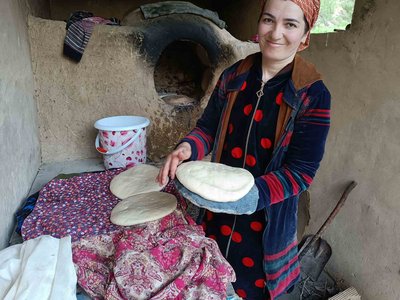 In einer schmalen Küche mit Lehmwänden präsentiert eine lächelnde Frau in usbekischer Kleidung eine Fladenbrot auf einem Teller.