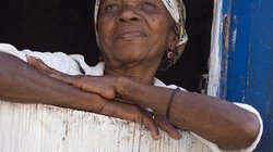 Ältere kapverdische Frau mit Kopftuch schaut über eine halbhohe weiße Tür in die Ferne, ihre Arme auf der Tür abgelegt