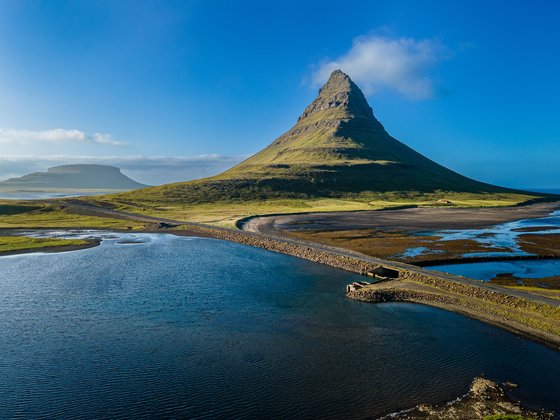 Die Snafellsnes-Halbinsel wird auch "Island in Miniatur" genannt Hinter einer Wasserfläche erhebt sich ein kegelförmiger Berg vor einem leuchtend blauen Himmel.