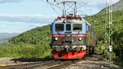 Man sieht ziemlich nah einen Triebwagen von vorne, der unten rot, in der Mitte schwarz und oben blau ist. Die Schiene verläuft nach vorne links aus dem Bild heraus. Den Zug umgibt eine bewaldete, hügelige Landschaft. Diagonal durchs Bild vom linken Bildmittelgrund bis in die obere rechte Ecke zieht sich ein hellblauer Himmel.