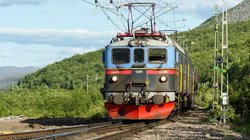 Man sieht ziemlich nah einen Triebwagen von vorne, der unten rot, in der Mitte schwarz und oben blau ist. Die Schiene verläuft nach vorne links aus dem Bild heraus. Den Zug umgibt eine bewaldete, hügelige Landschaft. Diagonal durchs Bild vom linken Bildmittelgrund bis in die obere rechte Ecke zieht sich ein hellblauer Himmel.