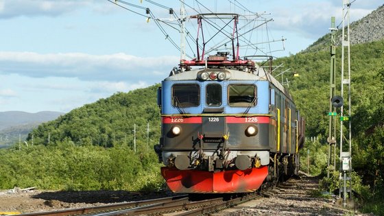 Man sieht ziemlich nah einen Triebwagen von vorne, der unten rot, in der Mitte schwarz und oben blau ist. Die Schiene verläuft nach vorne links aus dem Bild heraus. Den Zug umgibt eine bewaldete, hügelige Landschaft. Diagonal durchs Bild vom linken Bildmittelgrund bis in die obere rechte Ecke zieht sich ein hellblauer Himmel.