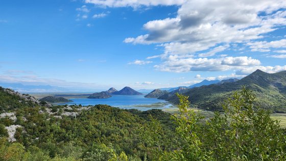 Von einem grünen Abhang blickt man weit in die grün-bewachsene Gebirgslandschaft. In der Bildmitte im Tal liegt eine große blaue Wasserfläche. Der Himmel ist blau mit vielen weißen Wolken in der rechten Bildhälfte.