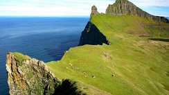 Aus dem blauen Meer links erhebt sich eine Steilklippe, die nach rechts abfällt und grün bewachsen ist.