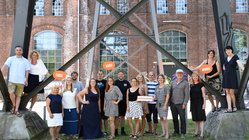 Teamfoto Reisen Mit Sinnen Das Reisen Mit Sinnen Team steht nebeneinander aufgestellt an einem Metallgestell vor einer Mauer.