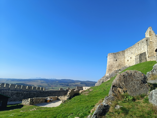Rechts im Bild erhebt sich auf einer Anhöhe eine massive Burgruine, links schaut man über die Umfassung der Burganlage tief in die flache Landschaft hinein. Die oberen zwei Drittel werden von blauem Himmel ausgefüllt abgesehen dort, wo rechts die Burg steht.