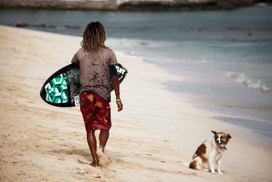 Ein Surfer am Strand Ein Surfer läuft am Strand entlang mit dem Surfbrett unter dem Arm. Daneben sitzt ein Hund.