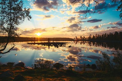 Im finnischen Sommer geht die Sonne kaum unter Eine Wasserfläche, um die Bäume stehen, wird von der milden Mitternachtssonne beschienen.