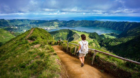 São Miguel: Eldorado für Wanderfreunde Auf einem Wanderpfad läuft eine Frau aus dem Bildvordergrund auf einen See zu, der sich im Bildhintergrund befindet.
