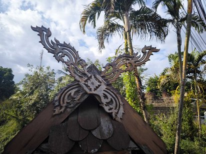 Man sieht einen mit Schnitzereien verzierten Dachgiebel. Hinter dem Haus ragen Palmen in den blauen Himmel.