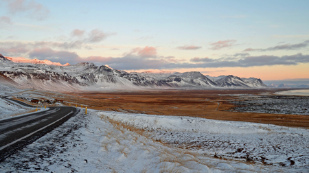 Snaefellsnes im Winter Man sieht eine leicht verschneite Landschaft vor einem hellblau-rosa dämmrigen Himmel, links durch Bild führt eine Straße.
