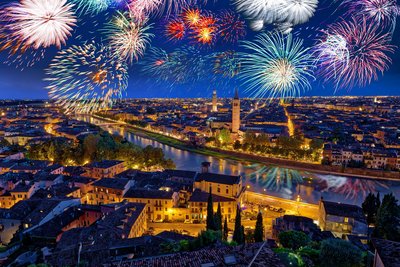 Aus der Vogelperspektive blickt man über das erleuchtete Häusermeer Veronas, das von der Etsch durchzogen wird. Am nächtlichen Himmel darüber erstrahlt ein Feuerwerk.