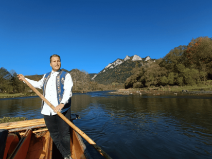 Links im Bild steht ein Flößer mit dem Stak-Ruder in der Hand, hinter und rechts neben ihm füllt Wasser das Bild aus bis zur Bildmitte horizontal, dahinter erheben sich Bäume, ein Hügel und viel blauer Himmel.