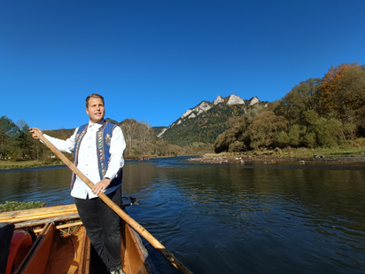 Links im Bild steht ein Flößer mit dem Stak-Ruder in der Hand, hinter und rechts neben ihm füllt Wasser das Bild aus bis zur Bildmitte horizontal, dahinter erheben sich Bäume, ein Hügel und viel blauer Himmel.