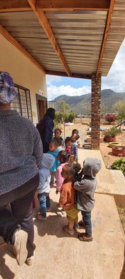 Vor einem hellen Gebäude unter einem Vordach steht eine Gruppe schwarzer Kindergartenkinder, zwei Erwachsene sind auch dabei.