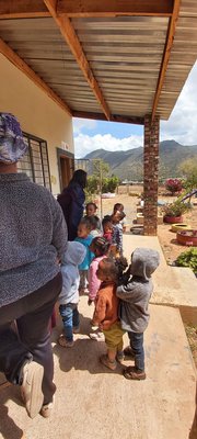 Vor einem hellen Gebäude unter einem Vordach steht eine Gruppe schwarzer Kindergartenkinder, zwei Erwachsene sind auch dabei.