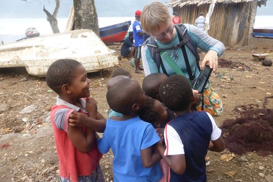 Eine blonde Europäerin mit Brille zweigt einer Gruppe afrikanischer Kinder ein Foto auf ihrem Handy. Im Hintergrund befinden sich mehrere kleine Boote am Ufer und eine Bambushütte.