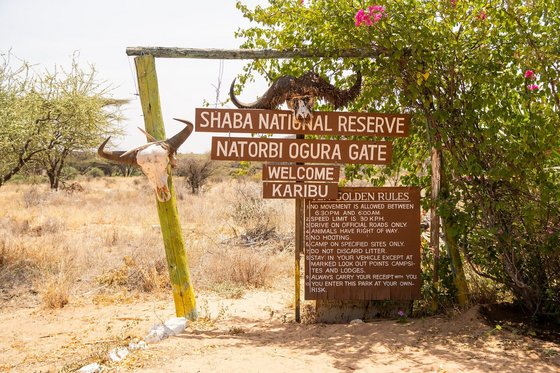Ein Schild mit Verhaltensregeln und dem Namen des Shaba National Reserves steht im Savannenboden und zeigt den Eingang des Nationalparks.