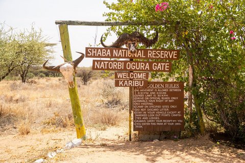 Ein Schild mit Verhaltensregeln und dem Namen des Shaba National Reserves steht im Savannenboden und zeigt den Eingang des Nationalparks.