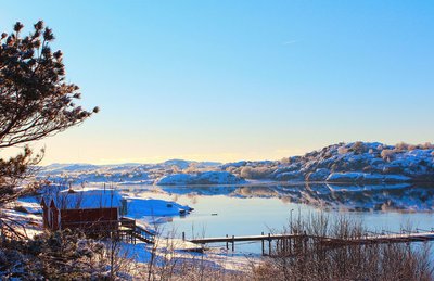 Rechts im Bild ist eine Wasserfläche, an deren Ufer links im Bild ein rotes Holzhaus steht. Die umgebende hügelige Landschaft ist weiß verschneit. Die obere Bildhälfte wird fast ausschließlich nur von hellblauem Himmel eingenommen.