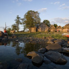 Eine kleine Gruppe von Touristen steht vor traditionellen Holzhäusern, am Ufer eines kleinen Bachlaufs