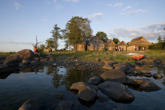 Eine kleine Gruppe von Touristen steht vor traditionellen Holzhäusern, am Ufer eines kleinen Bachlaufs