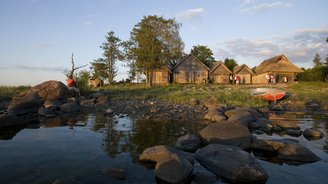 Eine kleine Gruppe von Touristen steht vor traditionellen Holzhäusern, am Ufer eines kleinen Bachlaufs