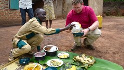 Ein hockender Afrikaner füllt einem daneben hockenden Weißen etwas zu Essen in eine Schüssel.