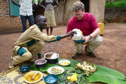 Vegetarisch unterwegs in Afrika Ein hockender Afrikaner füllt einem daneben hockenden Weißen etwas zu Essen in eine Schüssel.