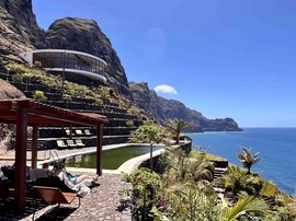 Foto eines Pools inmitten von grünen Sträuchern. Im Hintergrund sind hohe grüne Berge und das dunkelblaue Meer.