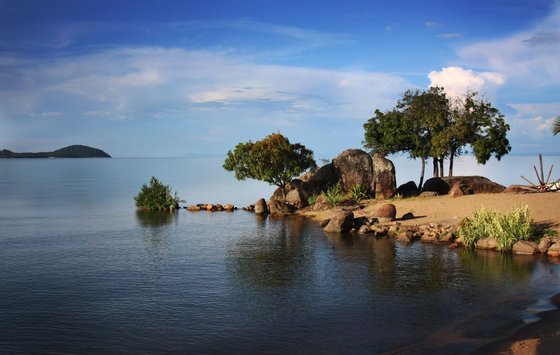 Ein Blick auf den Malawisee (Njassasee) mit klarem, schimmerndem Wasser, umgeben von einer vielfältigen Naturkulisse. Sanfte Hügel und grüne Uferlandschaften bilden einen malerischen Kontrast zur Weite des Sees. Die idyllische Umgebung lädt zum Entspannen und Erkunden ein.