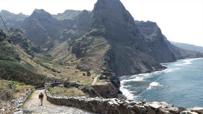 Ein einzelner Wanderer beschreitet vom Betrachter weg einen Küstenweg auf der kapverdischen "Wanderinsel" Santo Antao. Das Meer befindet sich auf der rechten Seite. Im Hintergrund erheben sich die Klippen und Berge.