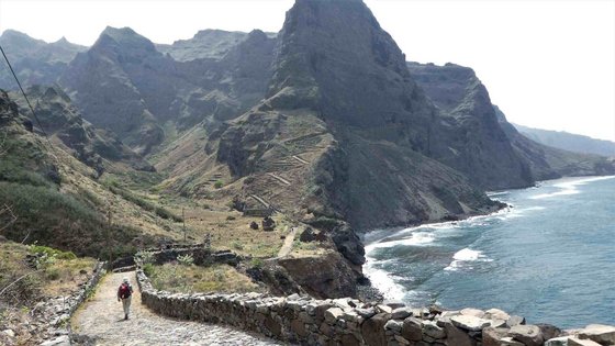 Ein einzelner Wanderer beschreitet vom Betrachter weg einen Küstenweg auf der kapverdischen "Wanderinsel" Santo Antao. Das Meer befindet sich auf der rechten Seite. Im Hintergrund erheben sich die Klippen und Berge.