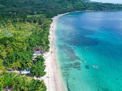 Eine Luftaufnahme zeigt einen langen, weißen Sandstrand auf der Insel Lombok, Indonesien. Links davon befinden sich bewachsene Palmenhaine und rechts das tiefblaue Meer.
