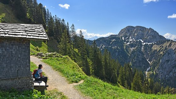Eine Frau sitzt auf einer Bank an einer Berghätte und blickt auf die grünen Berge.